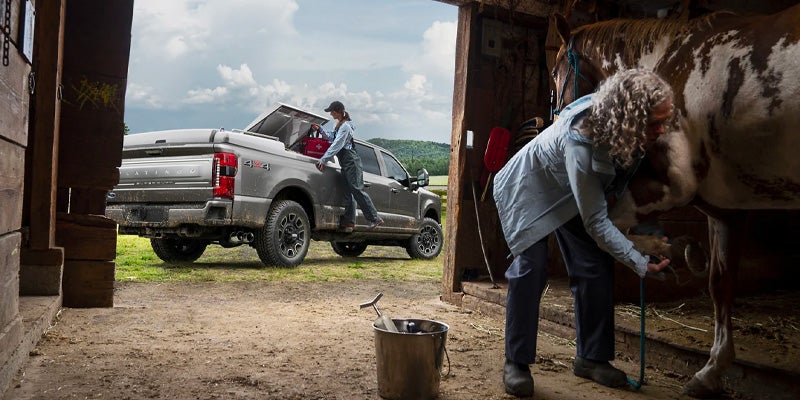 Pickup truck at barn, woman unloading gear, another grooming horse.